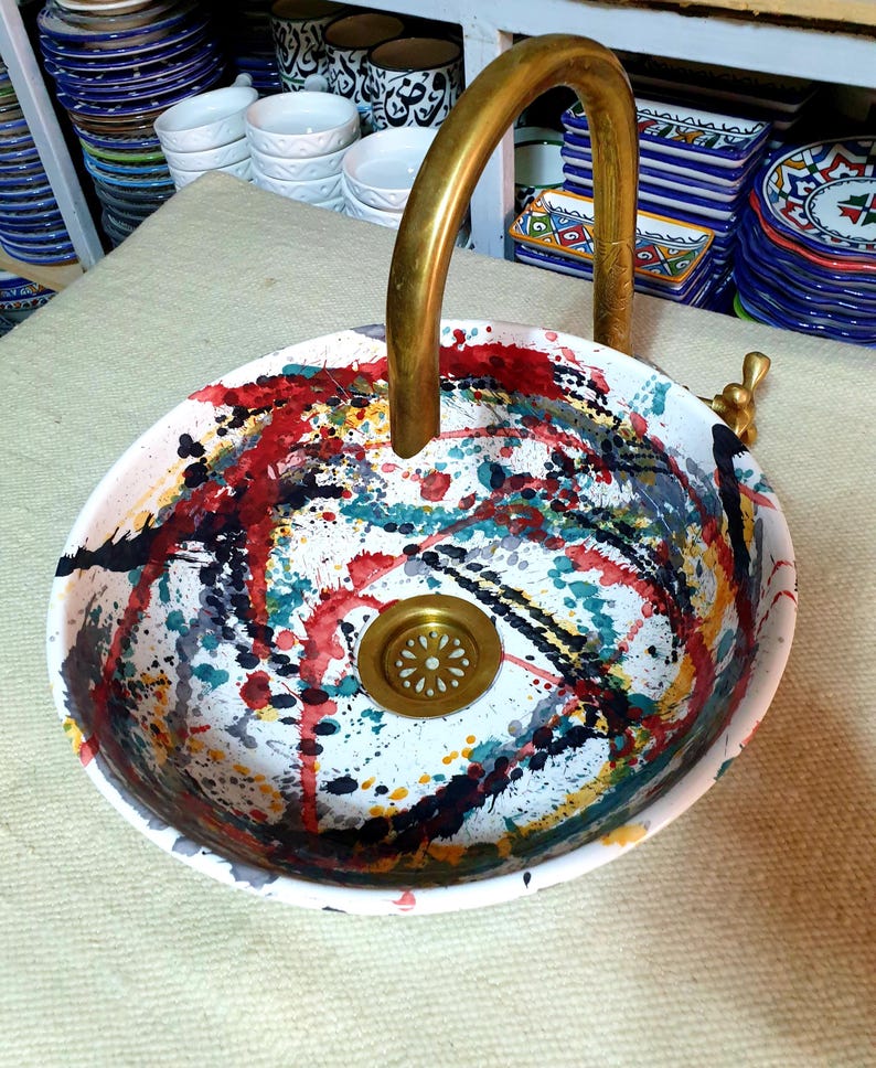 May include: A white ceramic sink with a gold faucet and a colorful abstract design. The sink has a drain in the center and is decorated with splashes of red, blue, green, yellow, and black paint.