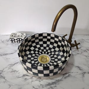 May include: A black and white checkered ceramic sink with a brass faucet. The sink has a circular design with a gold drain. A matching checkered soap dish is to the left.