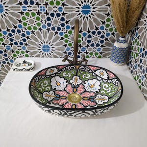May include: A round ceramic vessel sink featuring a hand-painted floral pattern in red, green, and black on a white background. The sink includes a brass-toned drain and is paired with a matching soap dish and a decorative vase with dried grass.