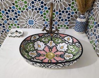 May include: A round ceramic vessel sink featuring a hand-painted floral pattern in red, green, and black on a white background. The sink includes a brass-toned drain and is paired with a matching soap dish and a decorative vase with dried grass.