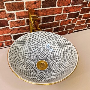 May include: A white ceramic sink with a black and white geometric pattern and a gold rim. The sink has a gold drain.