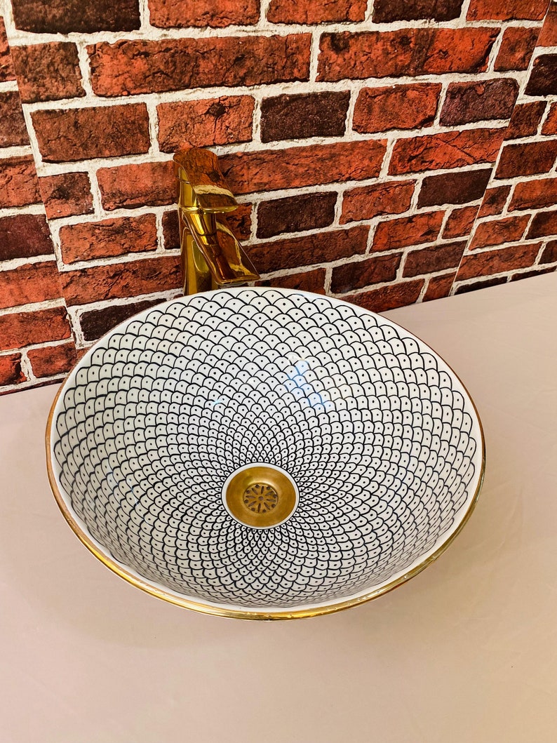 May include: A white ceramic sink with a black and white geometric pattern and a gold rim. The sink has a gold drain.