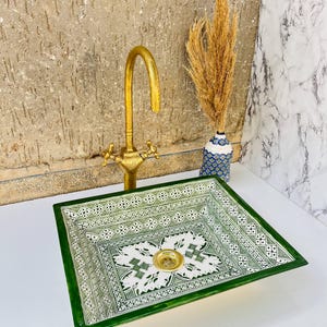 Może przedstawiać: Kwadratowy, zielono-biały ceramiczny zlew z misternym geometrycznym wzorem. Złoty kran jest zamontowany nad zlewem. Wazon z suszonymi roślinami stoi po prawej stronie zlewu. Zlew ma złoty odpływ.