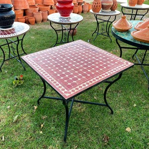 Peut inclure: Une table carrée avec un plateau en mosaïque géométrique rouge et blanc et un cadre en métal noir à quatre pieds incurvés. La surface de la table présente un motif répétitif d'octogones rouges entourés de bordures blanches, sur une pelouse.