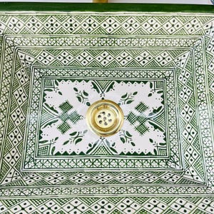 Może przedstawiać: Kwadratowa ceramiczna umywalka z białym i zielonym geometrycznym wzorem. Umywalka ma mosiężny odpływ i zieloną obw&oacute;dkę. Projekt zawiera motywy diament&oacute;w i kwiat&oacute;w. Umywalka jest prawdopodobnie do łazienki.