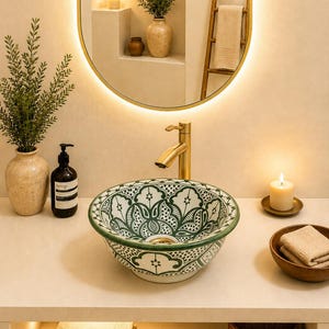 Könnte beinhalten: Ein rundes Aufsatzwaschbecken aus Keramik mit weißem Untergrund und komplexen grünen Blumen- und Geometriemustern. Das Waschbecken ist mit einer Armatur in gebürstetem Gold kombiniert und steht auf einer hellen Ablage mit goldenem Spiegel.