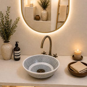 May include: A round ceramic vessel sink featuring a black and white radial striped pattern, paired with an antique brass gooseneck tap. The sink sits on a light-coloured countertop alongside a soap dispenser, a candle, and a wooden bowl with towels.