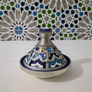 以下が含まれることがあります： 銀色の金属製のベースと蓋が付いた、装飾的なセラミック製のタジン鍋。蓋には青と白のセラミックノブが付いています。鍋は、銀色のアクセントが付いた青と白の幾何学模様が特徴です。