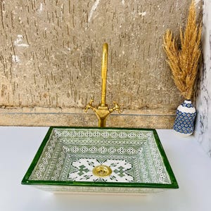 Może przedstawiać: Kwadratowa ceramiczna umywalka z zielono-białym geometrycznym wzorem. Umywalka ma złoty kran i złoty odpływ. Mały wazon z suszonymi roślinami znajduje się po prawej stronie umywalki.