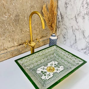 Może przedstawiać: Kwadratowy ceramiczny zlew z zielono-białym geometrycznym wzorem, połączony ze złotą baterią. Zlew posiada złoty odpływ. W tle wazon z suszonymi roślinami.