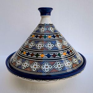 Puede incluir: Una olla de tagine de cerámica con tapa cónica y base ancha y poco profunda. La olla es principalmente blanca con intrincados patrones geométricos en azul, amarillo, verde y rojo. El borde y la parte superior de la tapa son de color azul oscuro.