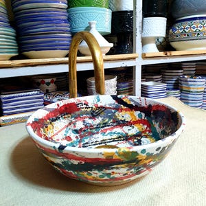 May include: A white ceramic sink with a colorful abstract design. The sink has splashes of red, yellow, blue, green, and black paint. A gold faucet is attached to the sink.