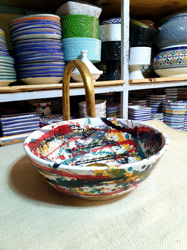 May include: A white ceramic sink with a colorful abstract design. The sink has splashes of red, yellow, blue, green, and black paint. A gold faucet is attached to the sink.