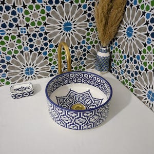 May include: A round, white ceramic sink with a blue geometric pattern. The sink has a gold faucet and is next to a small soap dish with a matching design. A vase with dried plants sits nearby, against a patterned tile wall.