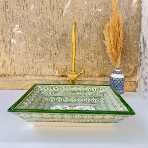 Może przedstawiać: Prostokątny ceramiczny zlew z zielono-białym geometrycznym wzorem. Zlew ma złoty kran i jest ustawiony na tle teksturowanej ściany. Po prawej stronie znajduje się mały wazon z suszonymi kwiatami.