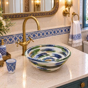 Peut inclure: Une vasque ronde en céramique ornée d'un motif abstrait peint à la main avec des coups de pinceau bleus et verts sur fond blanc, posée sur un comptoir beige à côté d'un robinet en laiton et d'accessoires de salle de bain assortis.