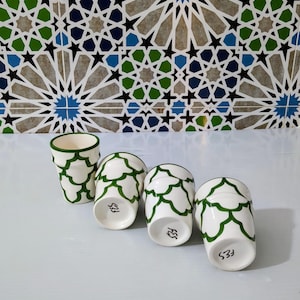 Puede incluir: Cuatro tazas de cerámica blanca con un patrón de hojas verdes y ribete verde. Las tazas están dispuestas en fila, con los fondos de las tazas visibles, cada una marcada con el texto "FES" y un número. El fondo presenta un diseño de azulejos geométricos.