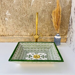 Może przedstawiać: Kwadratowa ceramiczna umywalka z zielono-białym geometrycznym wzorem. Umywalka ma mosiężną baterię i złoty odpływ. Wazon z suszonymi kwiatami stoi po prawej stronie umywalki. Umywalka stoi na białej powierzchni.