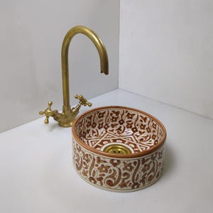 Op de afbeelding: Een ronde keramische wastafel met een bruin en wit bloemmotief. De wastafel wordt gecombineerd met een messing kraan. De kraan heeft twee kruisvormige handgrepen. De wastafel is een decoratief badkamerarmatuur.