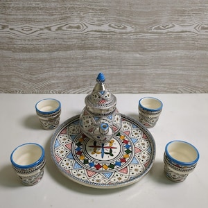 Op de afbeelding: Een Marokkaanse theeset met een zilveren theepot, een decoratieve schotel en vier kleine kopjes. De schotel en kopjes hebben een kleurrijk geometrisch ontwerp met blauwe, rode, gele en groene accenten. De theepot heeft een blauwe knop.