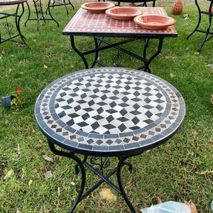 Peut inclure: Une table d'appoint ronde dotée d'une structure en métal noir avec des pieds incurvés et un plateau orné d'un motif mosaïque géométrique en carreaux noirs, blancs et gris. La table présente une bordure circulaire de petits carreaux en losange.