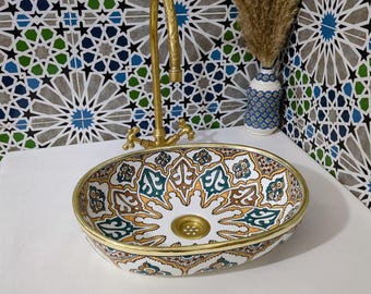 Può includere: Un lavabo ovale in ceramica con bordo dorato e motivi intricati in verde acqua, marrone e oro. Un rubinetto dorato è montato sopra il lavabo. Lo sfondo presenta una parete piastrellata a motivi con forme geometriche blu, verdi e grigie.