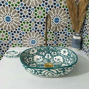 May include: A round ceramic vessel sink featuring a white base with intricate dark green floral and geometric patterns. The sink includes a gold-toned drain, a tall curved brass faucet, and a matching small rectangular soap dish.