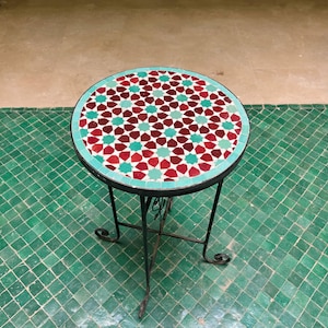 Può includere: Un tavolo rotondo con un piano in mosaico caratterizzato da un motivo geometrico di piastrelle rosse, bianche e turchesi. Il tavolo è sostenuto da una struttura in metallo nero con quattro gambe sottili e curve unite da decorazioni a voluta.