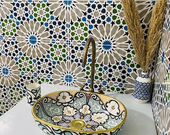 Puede incluir: Un lavabo ovalado de cerámica blanca con un diseño floral colorido. El lavabo tiene un borde dorado y un desagüe en el centro. El lavabo está colocado sobre un fondo de azulejos con patrones azules, verdes y blancos.