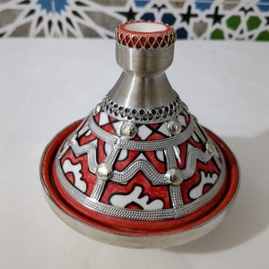 May include: A decorative tagine pot with a conical lid and a round base. The pot features a red and white geometric design with silver accents. The rim and base are silver. The top of the lid is red.