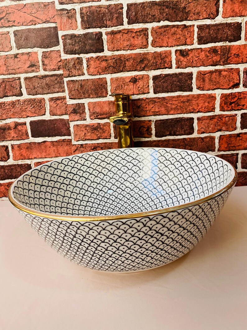 May include: A white ceramic sink with a black and white geometric pattern and a gold rim. The sink is round and has a slight curve to the bottom.
