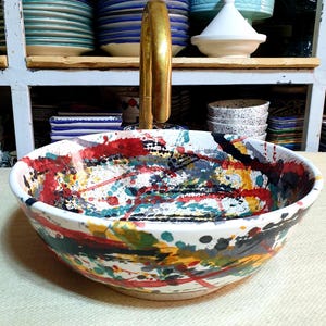 May include: A white ceramic sink with a colorful abstract design. The sink has splashes of red, yellow, blue, green, and black paint. The sink is on a white countertop.