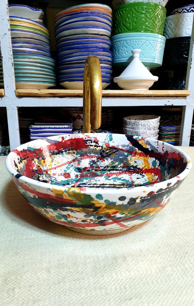 May include: A white ceramic sink with a colorful abstract design. The sink has splashes of red, yellow, blue, green, and black paint. The sink is on a white countertop.