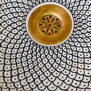 May include: A white ceramic sink with a black and white geometric pattern. The sink has a gold-coloured drain cover.