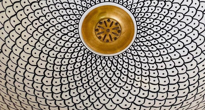 May include: A white ceramic sink with a black and white geometric pattern. The sink has a gold-coloured drain cover.