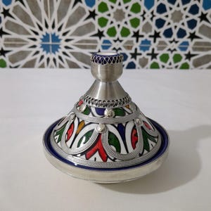 May include: A decorative, silver-toned tagine pot with a conical lid and a round base. The lid features colorful enamel designs in red, green, and blue, with silver filigree accents. The base is trimmed with a blue border.