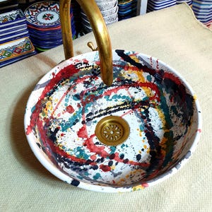 May include: A white ceramic sink with a brass faucet and a colorful abstract design. The sink has a round shape and a drain in the center. The design features splashes of red, yellow, blue, green, and black paint.