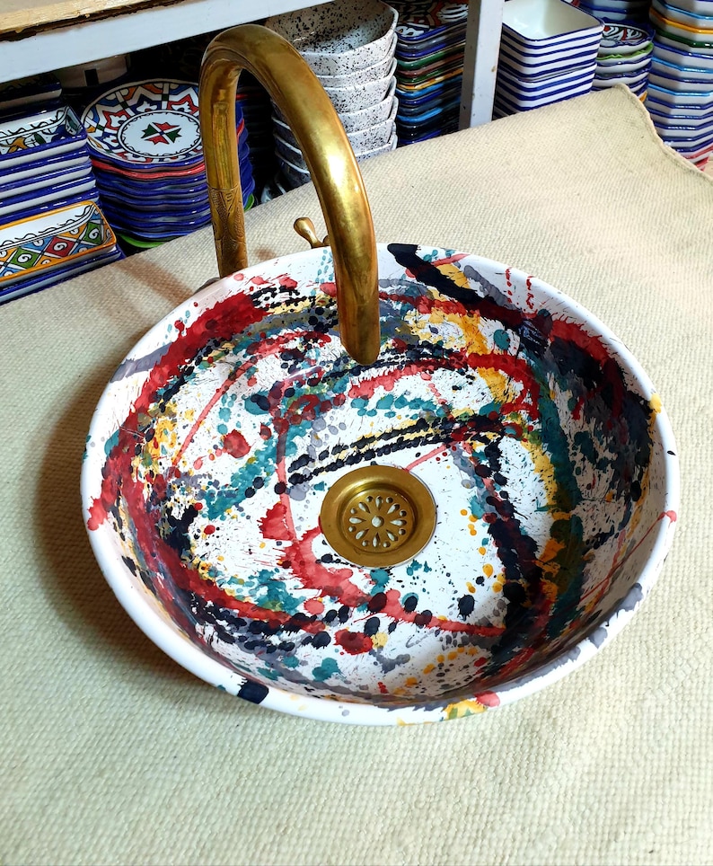 May include: A white ceramic sink with a brass faucet and a colorful abstract design. The sink has a round shape and a drain in the center. The design features splashes of red, yellow, blue, green, and black paint.