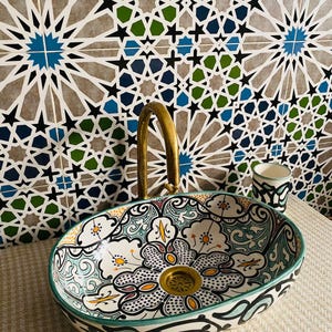 Puede incluir: Un lavabo ovalado de cerámica blanca con un diseño floral colorido. El lavabo tiene un desagüe de color dorado y está colocado sobre un fondo de azulejos geométricos azules, verdes y negros.