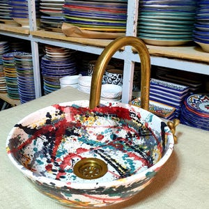 May include: A colorful ceramic sink with a gold faucet. The sink has a white base with splashes of red, yellow, blue, and green paint. The faucet is a simple, curved design.
