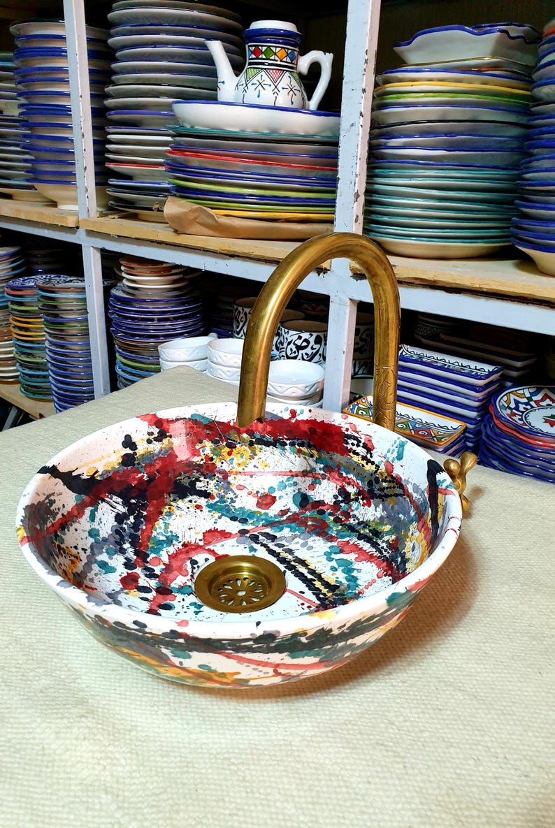 May include: A colorful ceramic sink with a gold faucet. The sink has a white base with splashes of red, yellow, blue, and green paint. The faucet is a simple, curved design.