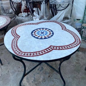 Peut inclure: Une table ronde avec un plateau en mosaïque présentant un fond blanc, un motif central en étoile bleu et rouge, et une bordure à motifs de losanges rouges. La table est soutenue par un cadre en métal noir avec quatre pieds incurvés.