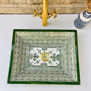Może przedstawiać: Kwadratowa ceramiczna umywalka z zielono-białym geometrycznym wzorem. Umywalka ma złoty odpływ i jest ustawiona na białej powierzchni. Złota bateria znajduje się nad umywalką, a w tle widoczny jest dekoracyjny wazon.