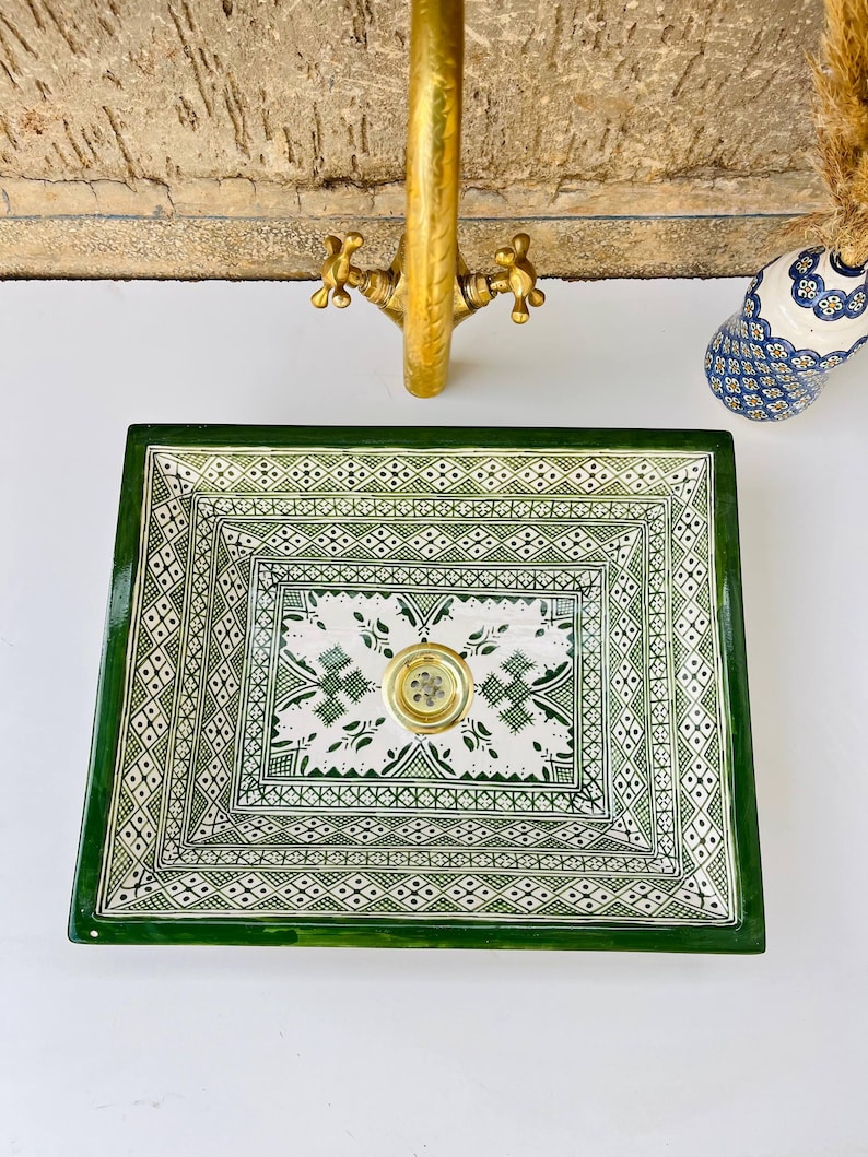 Może przedstawiać: Kwadratowa ceramiczna umywalka z zielono-białym geometrycznym wzorem. Umywalka ma złoty odpływ i jest ustawiona na białej powierzchni. Złota bateria znajduje się nad umywalką, a w tle widoczny jest dekoracyjny wazon.