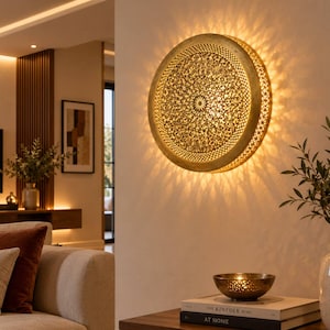 May include: A circular wall lamp with a gold-toned metal frame featuring an intricate, perforated floral pattern. The light emits a warm glow, casting detailed, star-like shadows onto the wall surface behind the fixture in a radial pattern.