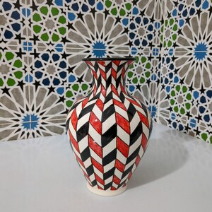 Könnte beinhalten: Eine dekorative Keramikvase mit einem geometrischen Muster aus roten, schwarzen und weißen Chevron-Mustern. Die Vase hat einen ausgestellten oberen Rand und einen weißen Sockel. Der Hintergrund zeigt eine gemusterte Fliesenwand mit blauen, grünen und grauen geometrischen Formen.