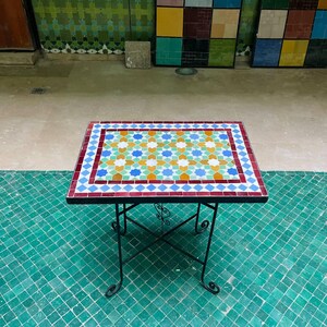 Puede incluir: Una mesa rectangular con un tablero de mosaico con un patrón geométrico en amarillo, azul, verde y blanco, bordeado por azulejos rojos y azules. La mesa se apoya en una estructura de metal negro con cuatro patas finas y curvas.