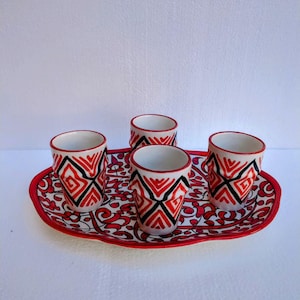 Può includere: Un set da tè in ceramica composto da quattro piccole tazze e un vassoio. Le tazze e il vassoio presentano un motivo geometrico rosso, nero e bianco. Il vassoio ovale ha un bordo rosso.