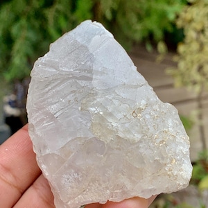 Può includere: Un cristallo di quarzo grezzo e traslucido con una superficie ruvida e irregolare. Il cristallo ha un colore bianco latte pallido con aree di trasparenza. La pietra naturale è tenuta in mano, con uno sfondo verde sfocato.