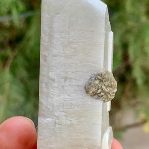 Puede incluir: Un espécimen mineral cristalino de color claro con una superficie rugosa y texturizada. Una pequeña formación mineral redonda de color beige está adherida al lado del cristal más grande. El cristal está sostenido en la mano de una persona.